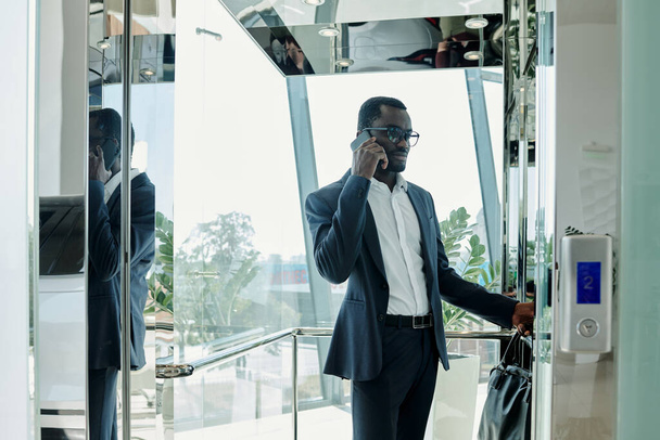 現代オフィスセンターの透明なエレベーターの中に立つスーツ姿の若いエレガントなビジネスマンと携帯電話で通信 - 写真・画像