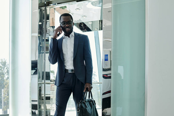Confident African American businessman in elegant suit talking on smartphone while going out of elevator with transparent walls - Photo, Image