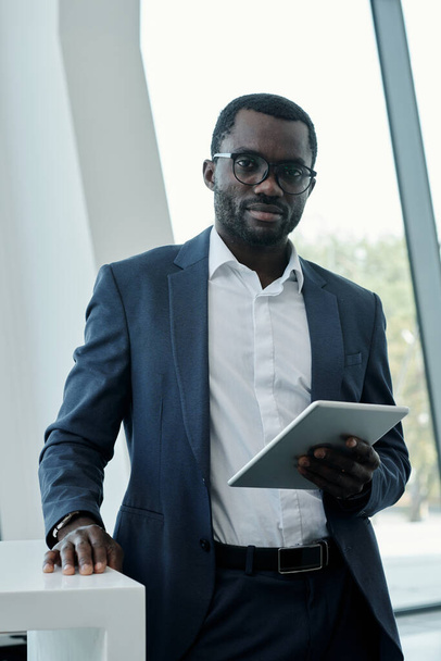 Young confident broker or chief executive officer in elegant suit using tablet while standing by workplace and looking at camera - Fotó, kép