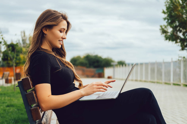 Außenporträt einer blonden jungen Frau, die an einem windigen Tag auf einer Bank sitzt und am Laptop arbeitet. - Foto, Bild