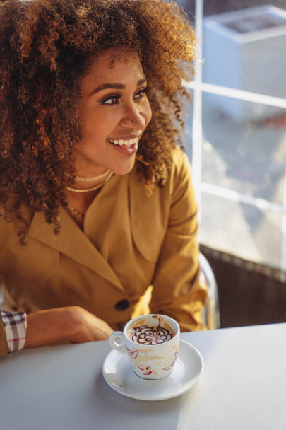 Афроамериканка, сидящая за столом в кафетерии с чашкой, поднятой и смотрящей в камеру - Фото, изображение