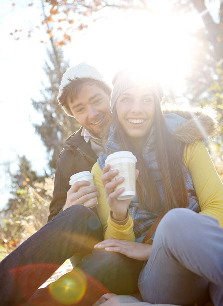 Part of their weekend routine- coffee in the park. An affectionate young couple sitting together while enjoying some coffee in the outdoors - Фото, изображение