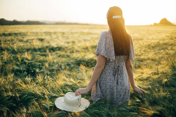 Piękna kobieta w kwiatowej sukience spacerująca po polu jęczmienia w świetle zachodu słońca. Atmosferyczny moment spokoju, rustykalne powolne życie. Stylowe kobiety gospodarstwa słomkowy kapelusz i korzystających wieczorem wsi letniej - Zdjęcie, obraz