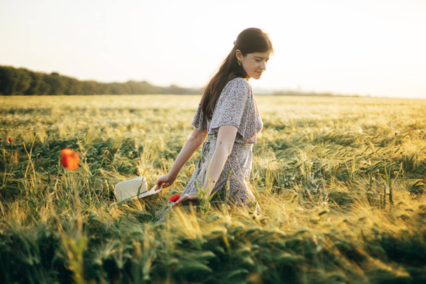 Beautiful woman in floral dress walking in barley field in sunset light. Atmospheric moment, rustic slow life. Stylish female holding straw hat and enjoying evening summer countryside - Foto, imagen
