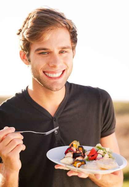 Portrait, smile and a man eating food outdoor on a summer day for health, diet or nutrition. Happy, morning and lifestyle with a handsome young person enjoying a fresh meal alone outside in nature. - Photo, Image