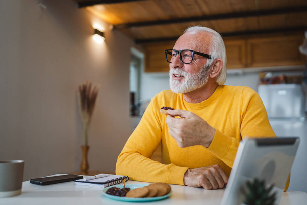 Ένας ηλικιωμένος άνδρας καυκάσιος γέρος συνταξιούχος κάθεται σε ένα τραπέζι στο σπίτι βάζοντας μαρμελάδα σε ένα μπισκότο που ετοιμάζεται να φάει πρωινό απολαύστε ένα σνακ χαλαρό στο περιβάλλον του σπιτιού του πραγματικό πρόσωπο αντιγραφή χώρου καθημερινή ρουτίνα - Φωτογραφία, εικόνα