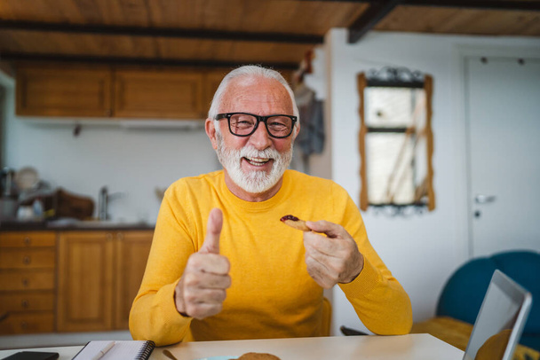 Un hombre mayor caucásico viejo pensionista masculino sentarse en una mesa en casa poniendo mermelada en una galleta preparándose para desayunar disfrutar de un aperitivo relajado en su hogar ambiente persona real copiar espacio rutina diaria - Foto, Imagen