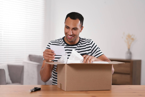 Happy young man opening parcel at table indoors. Internet shopping - Фото, зображення