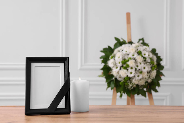 Photo frame with black ribbon, burning candle on table and wreath of flowers near white wall indoors, space for text. Funeral attributes - Photo, image