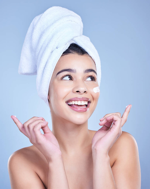 A beautiful smiling mixed race woman applying cream to her cheek. Hispanic model with towel wrapped hair using lotion against a blue copyspace background. - Foto, Imagen