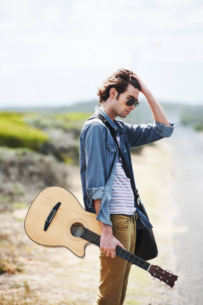 Ροκ εν ρολ στην άκρη του δρόμου. Trendy νεαρός άνδρας κρατώντας μια ακουστική κιθάρα στέκεται στην άκρη του δρόμου - Φωτογραφία, εικόνα