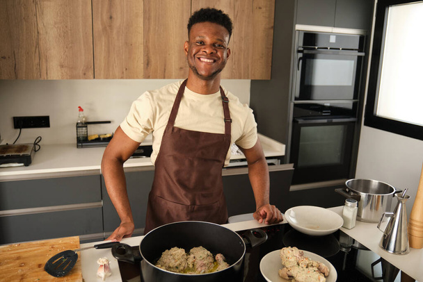 Νεαρός Αφρικανός άνδρας χαμογελά και κοιτάζει την κάμερα ενώ ετοιμάζει τηγανητό κοτόπουλο κιμά με λαχανικά συνταγή σε μια κουζίνα. - Φωτογραφία, εικόνα