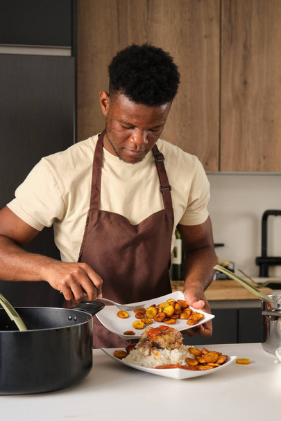 Jonge zwarte man serveert weegbree op tomatensaus gebakken kip gehakt en rijst plaat in een keuken. - Foto, afbeelding