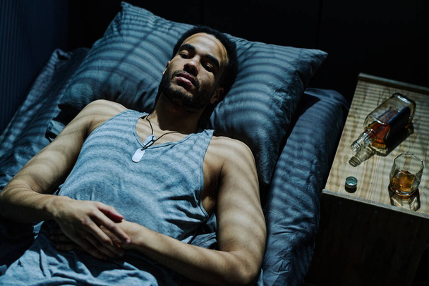 Вид зверху молодий чоловік лежить на ліжку з пляшкою віскі на нічному столі і страждає від безсоння після травматичної події
 - Фото, зображення