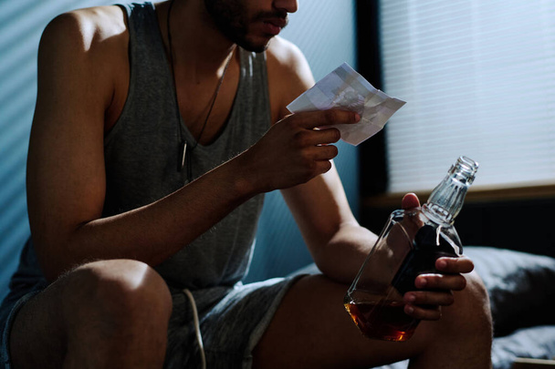 Νεαρός καταθλιπτικός άντρας με αλκοολούχο ποτό κοιτάζει φωτογραφία του με ποδήλατο ενώ πάσχει από μετατραυματικό σύνδρομο και αϋπνία - Φωτογραφία, εικόνα