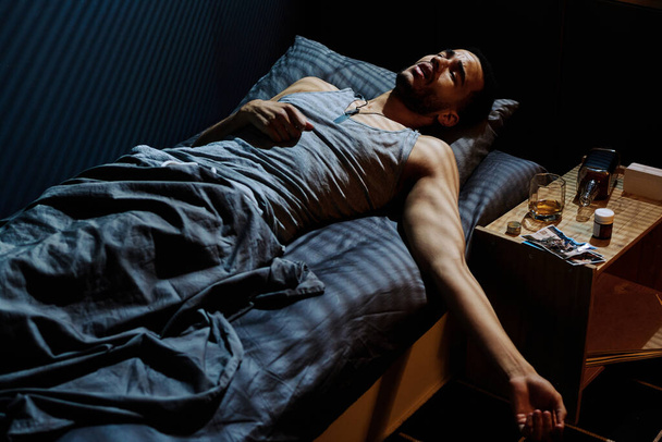 Gestresster junger Mann, der auf dem Nachttisch liegend mit Whisky in Flasche und Glas, Schlaftabletten und Fotos einzuschlafen versucht - Foto, Bild