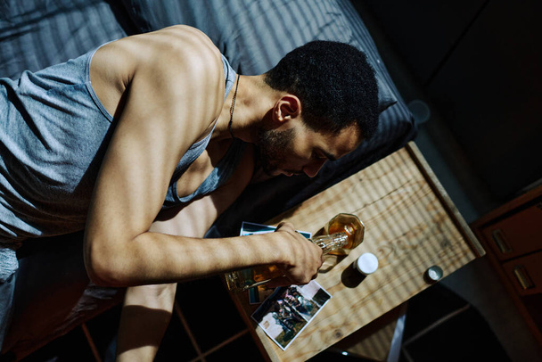 Boven schot van jonge man met post traumatisch syndroom gieten whisky in glas terwijl liggend op bed door kleine nachtkastje om middernacht - Foto, afbeelding