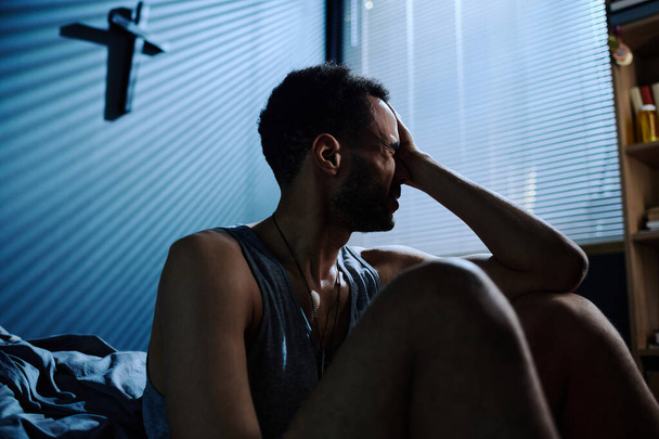 Młody zestresowany mężczyzna dotykający głowy siedząc przy łóżku po bezsennej nocy i czując się bezradny wobec konsekwencji PTSD - Zdjęcie, obraz