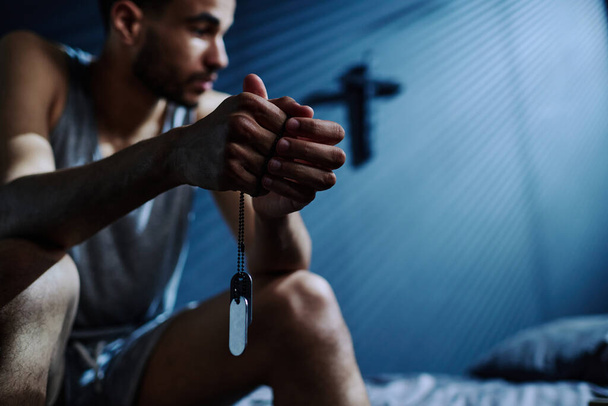 Handen van jonge slapeloze man die zilveren ketting met medaillons vasthoudt terwijl hij op bed zit na een slapeloze nacht en aan iets denkt. - Foto, afbeelding