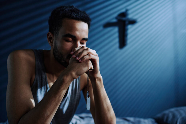 Jeune homme déprimé avec des médaillons métalliques sur la chaîne dans ses mains priant Dieu pour l'aider à s'endormir tout en étant assis sur le lit - Photo, image