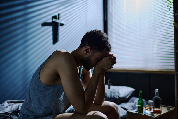 Boční pohled na mladého nekonejšivého muže s posttraumatickým syndromem, jak se ohýbá dopředu, zatímco sedí na posteli s rukama u obličeje - Fotografie, Obrázek