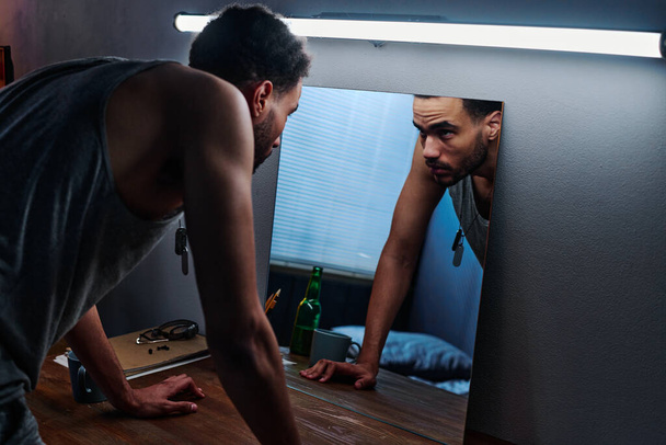 Jovem com transtorno pós-traumático dobrando-se sobre a mesa e olhando para seu reflexo no espelho enquanto sofre de insônia - Foto, Imagem