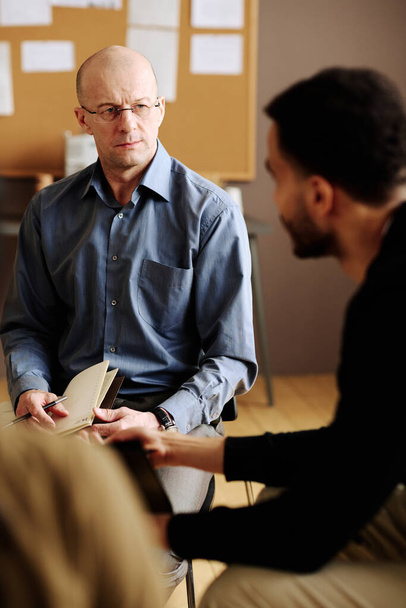 Confident mature counselor looking at one of patients with post traumatic disorder while listening to him during session - Photo, Image
