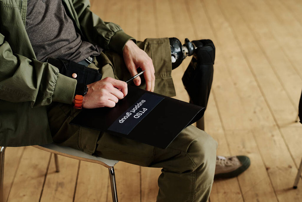 Young man with disability holding black folder of PTSD support group while sitting on chair during psychological session - Photo, Image