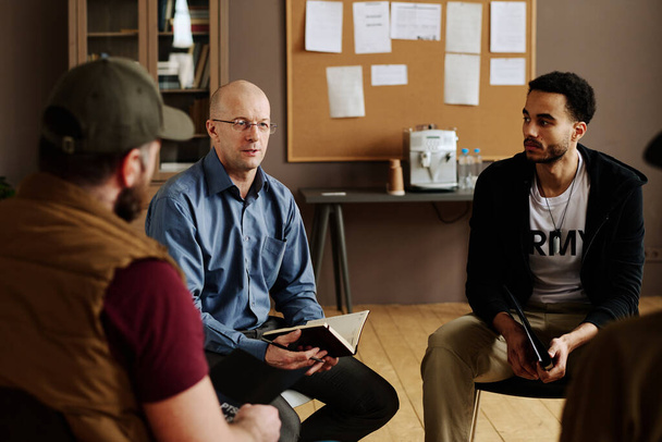 Уверенный взрослый консультант мужского пола консультирует людей с посттравматическим синдромом, сидящих вокруг него и слушающих его на сеансе - Фото, изображение