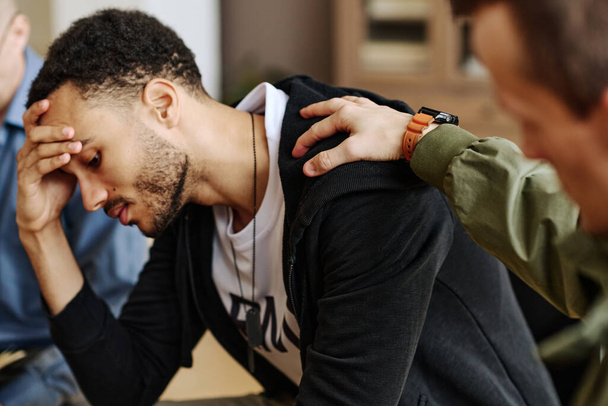 Junger gestresster Mann berührt Kopf, während einer der Begleiter die Hand auf seiner Schulter hält und ihn während der psychologischen Sitzung stützt - Foto, Bild