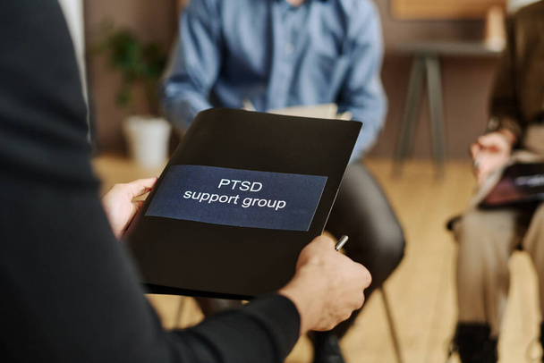 Selektywne skupienie się na czarnym folderze z naklejką mówiącą grupa wsparcia PTSD posiadana przez młodego pacjenta płci męskiej psychoterapeuty siedzącego z przodu - Zdjęcie, obraz