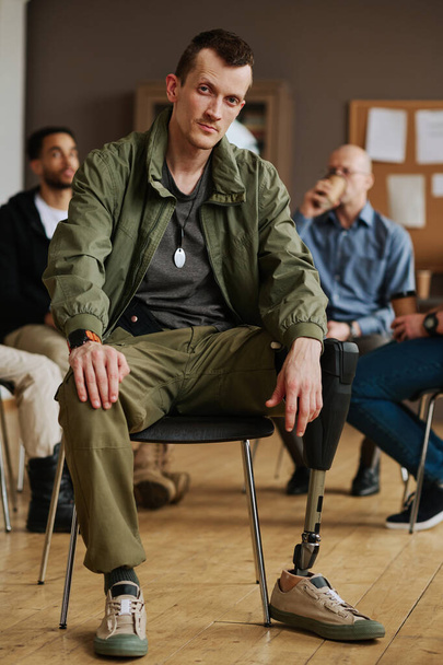 Young man with prosthetic leg sitting in front of camera against counselor and other attendants of PTSD support group having break - Photo, Image