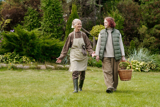 Старшая женщина в повседневной одежде разговаривает со своей внучкой, несущей корзину, в то время как обе движутся по зеленой лужайке против зеленой растительности - Фото, изображение