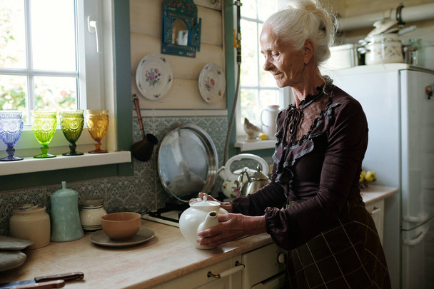 Heitere Seniorin hält Teekanne, während sie Tee für ihre Gäste aufbrüht oder morgens im Landhaus frühstückt - Foto, Bild