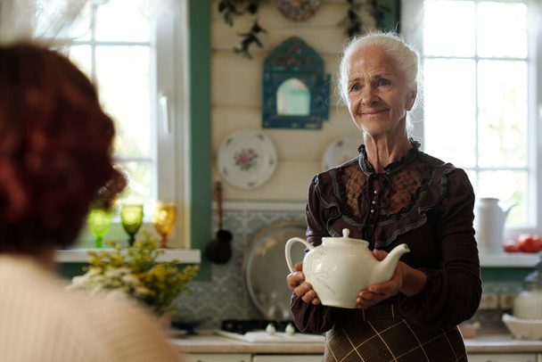 Happy grandmother with white porcelain teapot moving towards her granddaughter waiting for breakfast in the kitchen - Photo, image