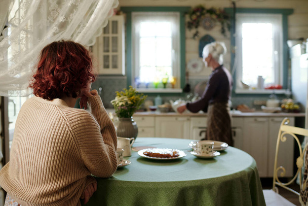 Visão traseira da menina em pulôver bege de malha sentada à mesa com torta caseira e esperando por sua avó preparando café da manhã - Foto, Imagem