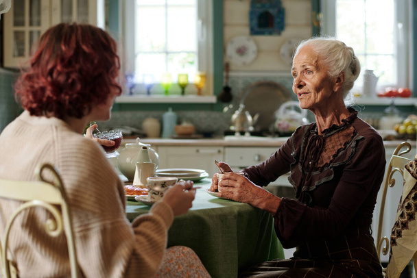 Idős nő alkalmi öltözetben teázik és beszél a fiatal unokájával, miközben mindketten a konyhában ülnek az asztalnál. - Fotó, kép