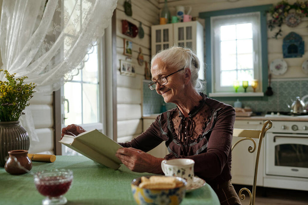 Mujer de edad feliz en ropa de casualwear inteligente y anteojos leer novela o libro de cuentos mientras está sentado junto a la mesa en la cocina de la casa de verano - Foto, imagen