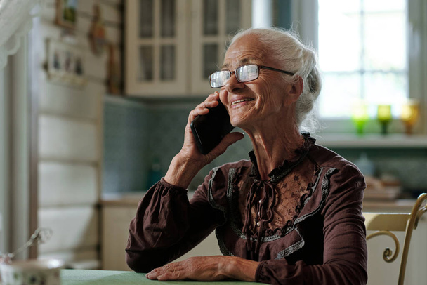 Mujer mayor alegre en gafas y blusa inteligente hablando con su familia en el teléfono móvil mientras está sentada junto a la mesa en la casa de campo - Foto, imagen