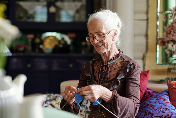 Senior smiling woman in casualwear knitting new clothes for her family members while sitting on couch in summer house at leisure - Photo, Image