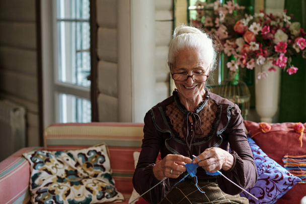 Ευτυχισμένη γιαγιά με κομψά ρούχα κάθεται στον καναπέ και πλέκει ζεστά μάλλινα χειμωνιάτικα ρούχα για τα μέλη της οικογένειάς της - Φωτογραφία, εικόνα