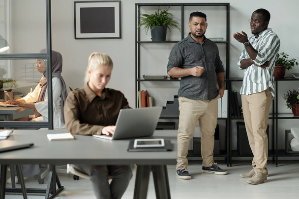 Dois homens interculturais em casualwear bullying mulher caucasiana loira com laptop sentado à mesa na frente deles e networking - Foto, Imagem
