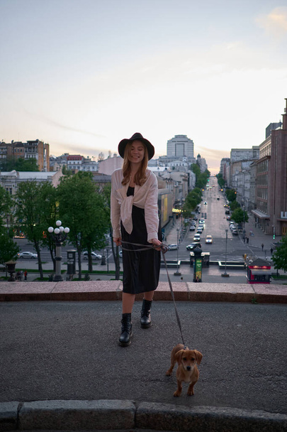 Fashion portrait of stylish elegant lady with a dog. Beautiful blonde young caucasian girl in black jacket and hat posing looking at camera with city view. Sunset time. High quality image - Photo, Image