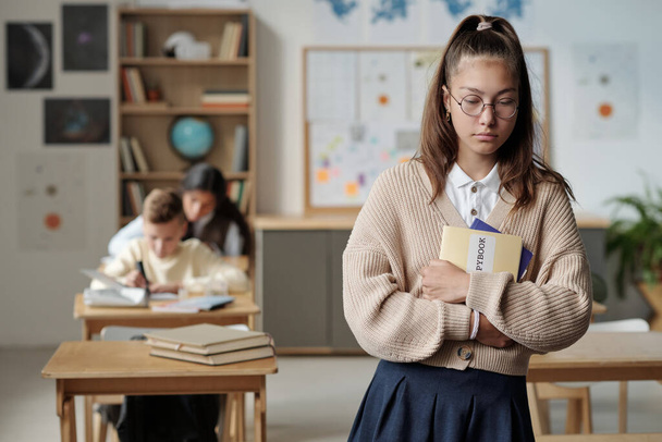 Upset and lonely schoolgirl with copybooks standing in front of camera or moving to her place on background of intercultural classmates - Photo, Image