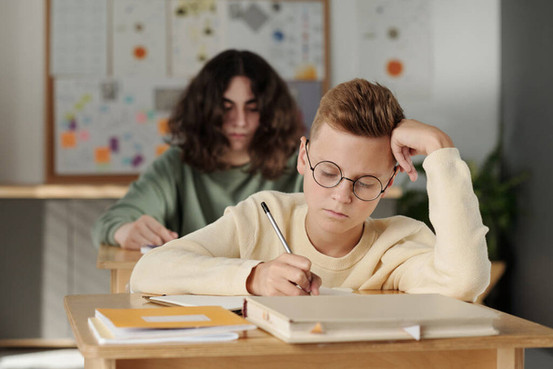 Slimme jeugdige schooljongen in brillenglazen die aantekeningen maakt in een copybook tijdens een individuele opdracht tijdens een les tegen zijn klasgenoot - Foto, afbeelding