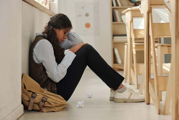 Side view of lonely and upset African American schoolgirl sitting on the floor of classroom while suffering from bullying of her classmates - Photo, Image