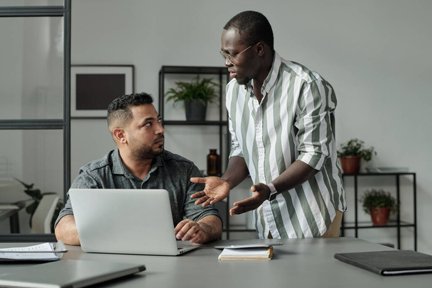 Młody, zirytowany afro-amerykański pracownik patrzący na wielonarodowego lub latynoskiego mężczyznę siedzącego przy biurku przed laptopem podczas rozmowy - Zdjęcie, obraz