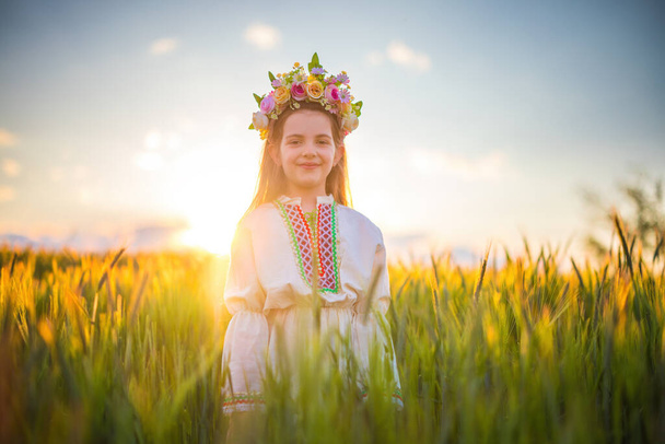 Beautiful young girl with flower chaplet, ethnic folklore dress with traditional Bulgarian embroidery during sunset on a wheat agricultural field - Фото, изображение