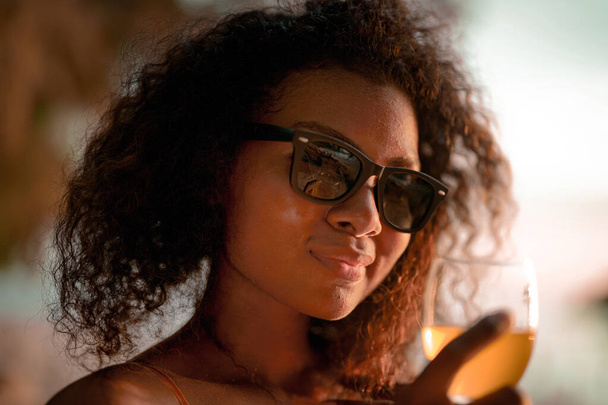 Africká žena pije koktejlový džus, když sedí na lavičce na pláži v tropické pláži. Mladý cestovatel ve žlutých bikinách a chladí krásu přírody. Wanderlust a pojetí cestování.  - Fotografie, Obrázek