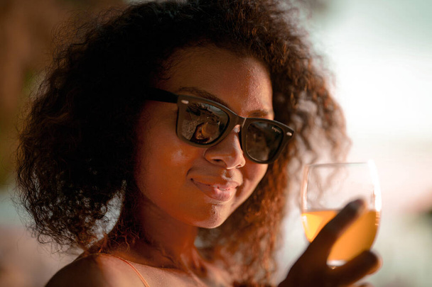 Αφρικανή γυναίκα πίνει χυμό κοκτέιλ όταν κάθεται στον πάγκο της παραλίας στην τροπική παραλία. Νεαρός ταξιδιώτης που φοράει κίτρινο μπικίνι και χαλαρώνει την ομορφιά της Φύσης. Wanderlust και ταξιδιωτική έννοια.  - Φωτογραφία, εικόνα